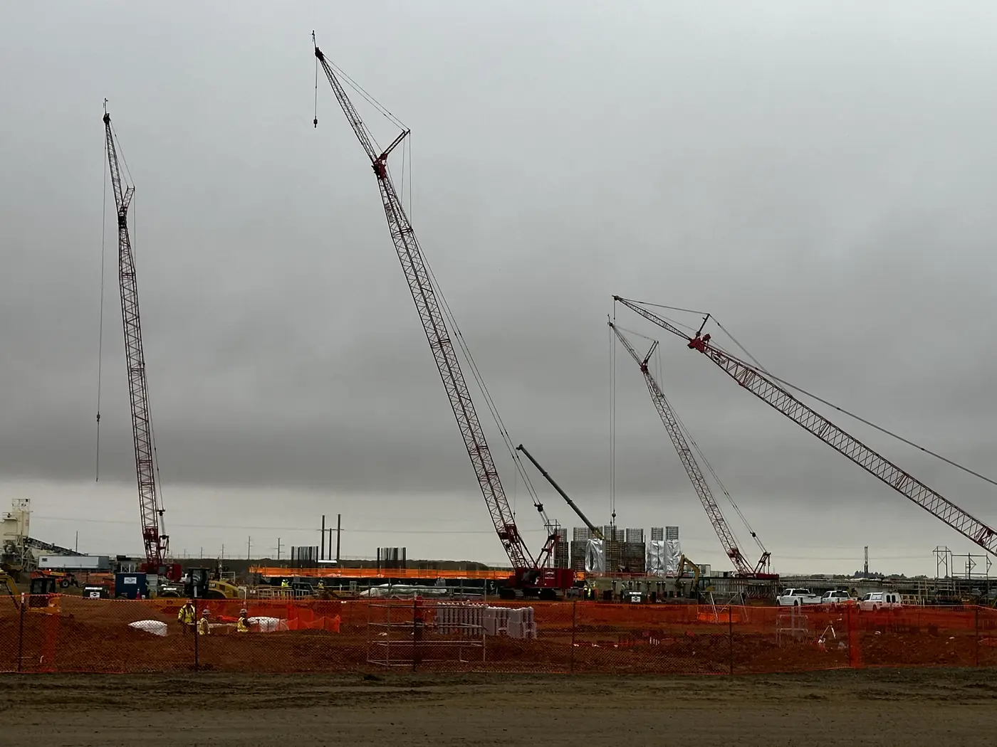 Three crawler cranes standing over a construction site behind an orange safety fence under a grey sky