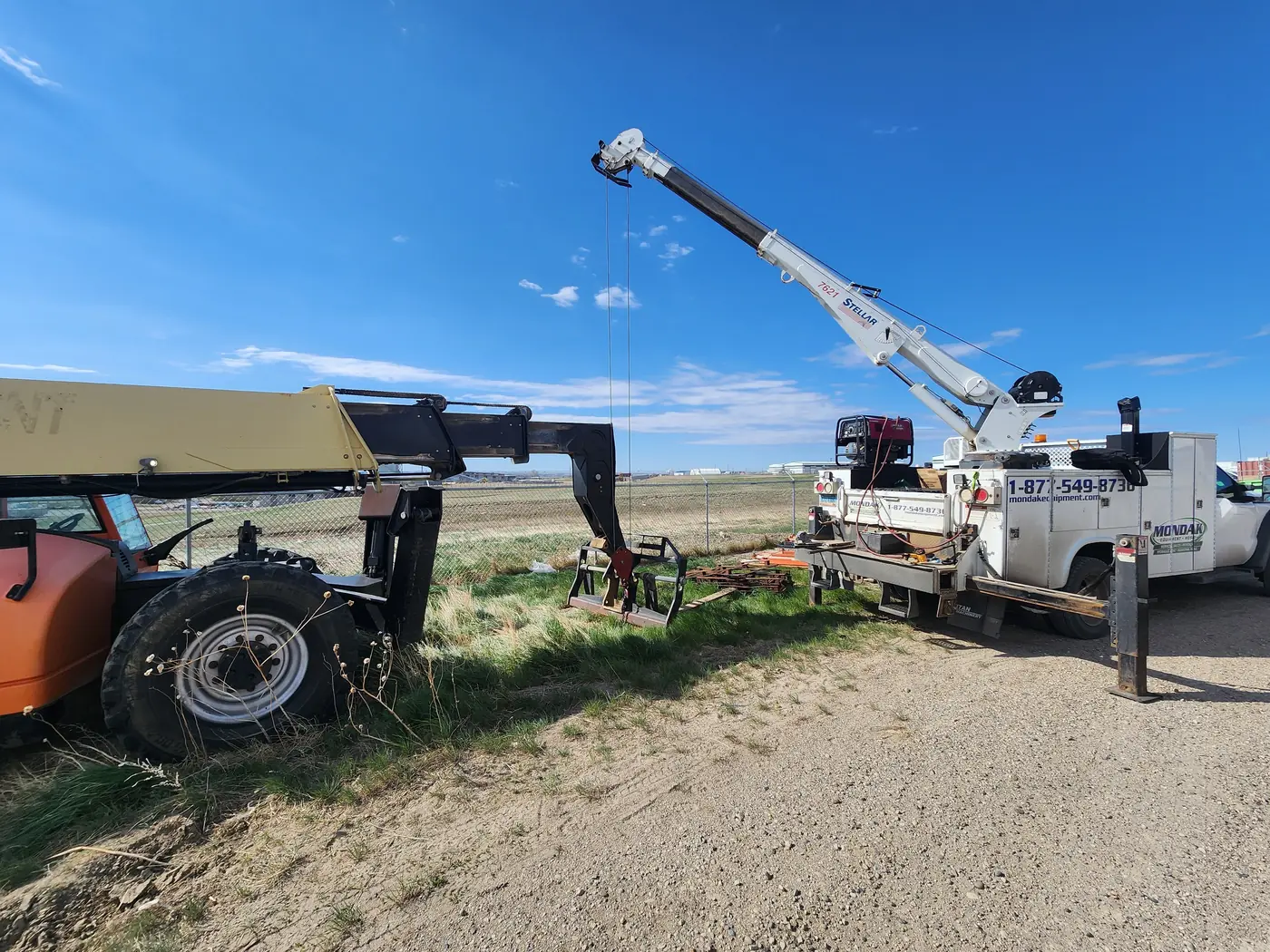 A JLG telehandler parked in the grass beside a fence with a Mondak service truck and its crane working alongside