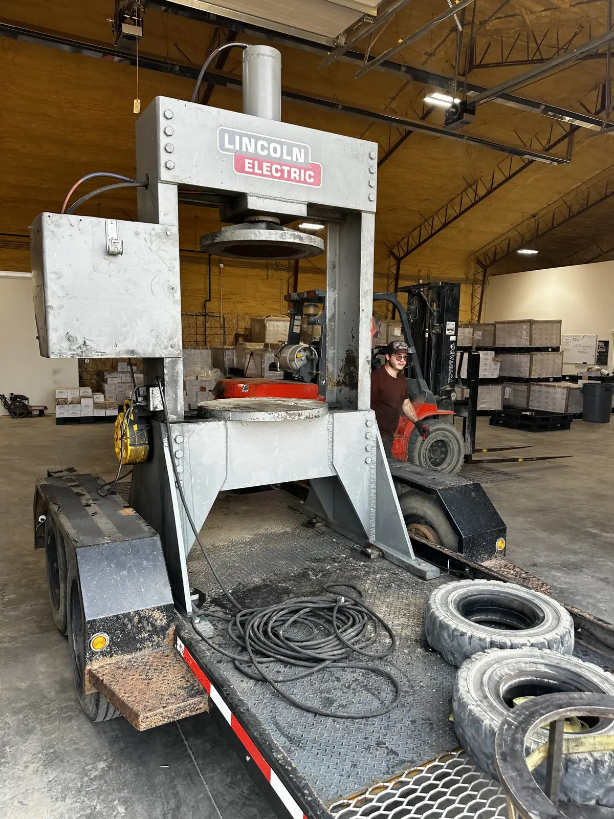 Trailer mounted tire press in a shop, with solid tires on the deck and a worker standing beside a forklift